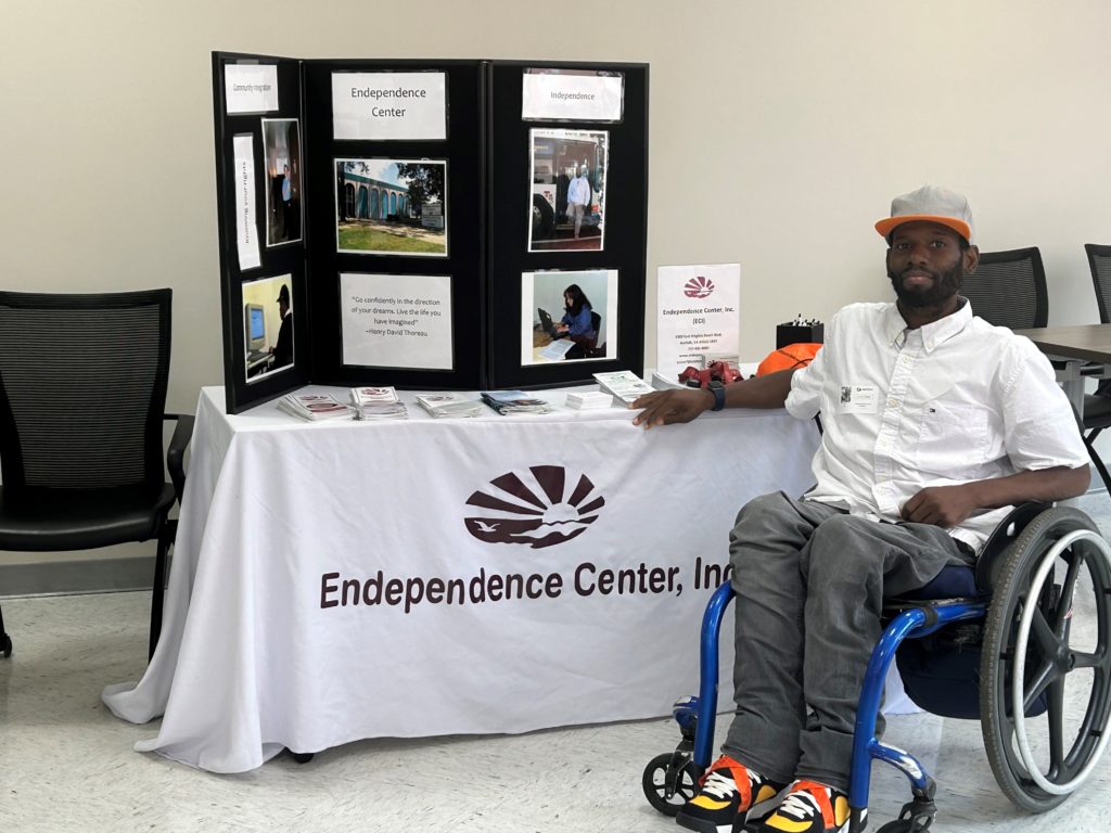 ECI staff member who is African American in a wheelchair wearing jeans and a white collars shirt is in front of ECI display table with a white table cloth with ECI logo and a black tri board which has information about ECI services
