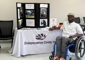 ECI staff member who is African American in a wheelchair wearing jeans and a white collars shirt is in front of ECI display table with a white table cloth with ECI logo and a black tri board which has information about ECI services