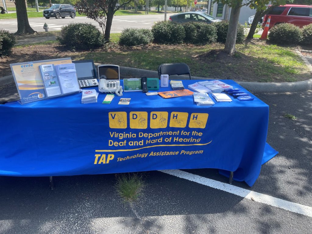 A table with a blue tablecloth and yellow lettering and logos. The tablecloth has "VDDHH" printed. On top of the table are brochures and TTY devices.