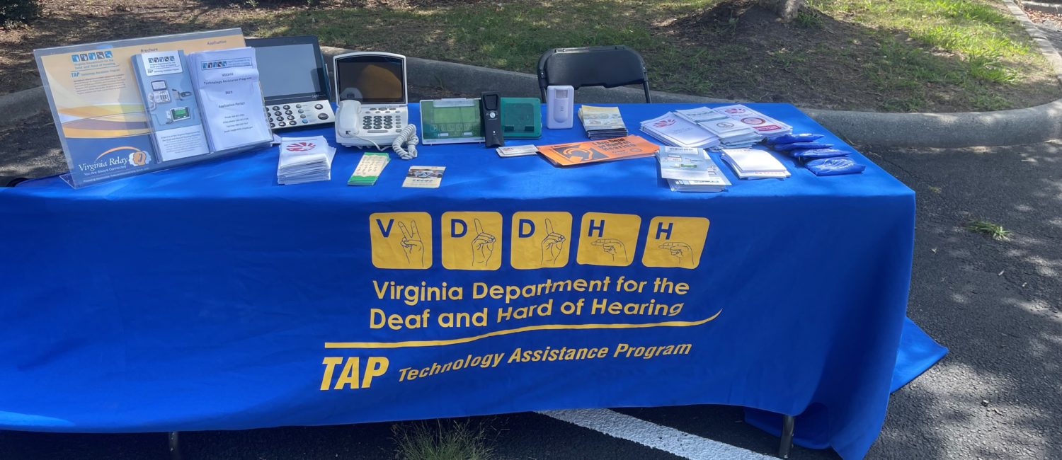 A table with a blue tablecloth and yellow lettering and logos. The tablecloth has "VDDHH" printed. On top of the table are brochures and TTY devices.