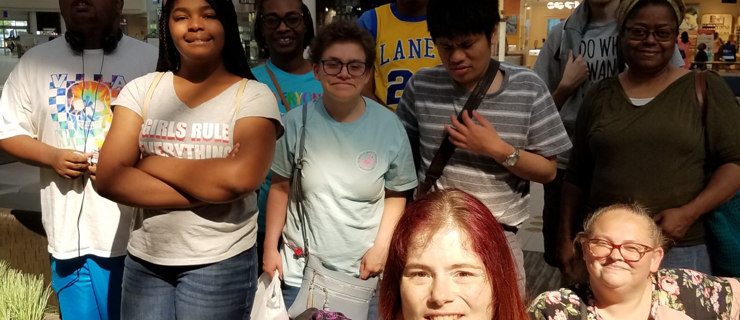 A Color picture of a female ECI staff member using a wheelchair. Behind her stands a group of teens of all ages with other ECI staff, taken at a community outing.