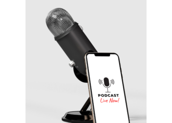 A black podcast microphone is on a desk. Leaning against the microphone is a cell phone, with the words "Podcast Live Now" on the screen.