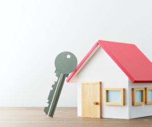 A house key is standing on end, leaning against a scale model of a house with a red roof.