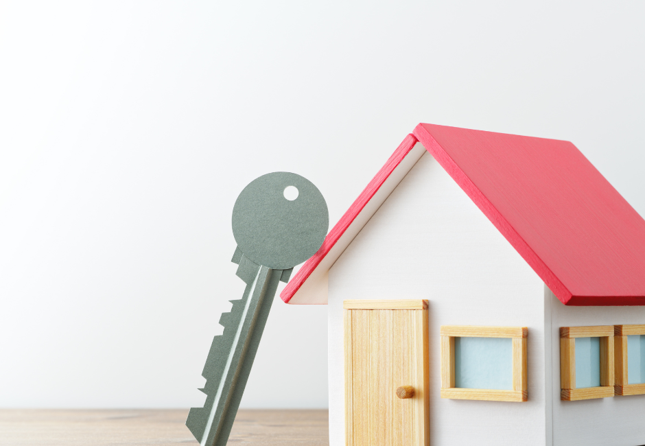 A house key is standing on end, leaning against a scale model of a house with a red roof.