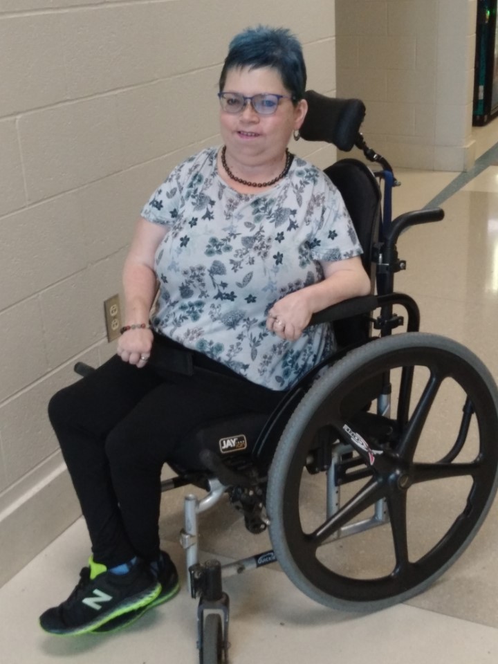 Picture of Lisbet Dula, who is a white woman using a wheelchair, wearing a floral print top, black pants, and green shoes. She is wearing glasses and has blue hair.