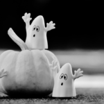 Two ghost figurines rest on a table and pumpkin, with their arms raised.