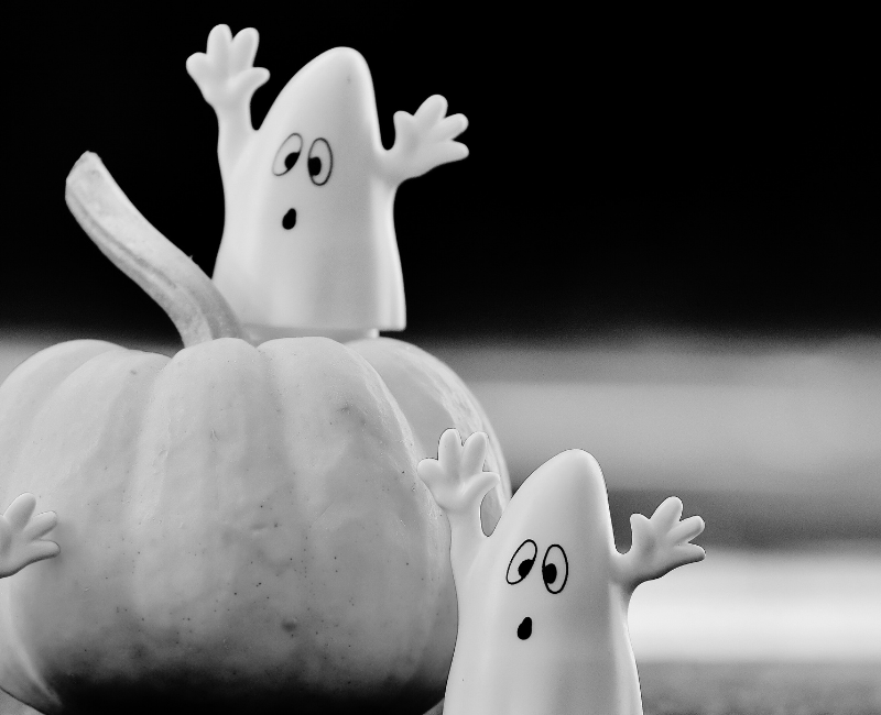 Two ghost figurines rest on a table and pumpkin, with their arms raised.