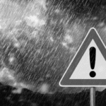 A triangular warning sign with a large exclamation mark in the center. The sign is set against a stormy background with heavy rain and dark clouds.