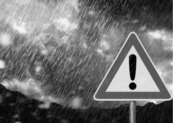 A triangular warning sign with a large exclamation mark in the center. The sign is set against a stormy background with heavy rain and dark clouds.